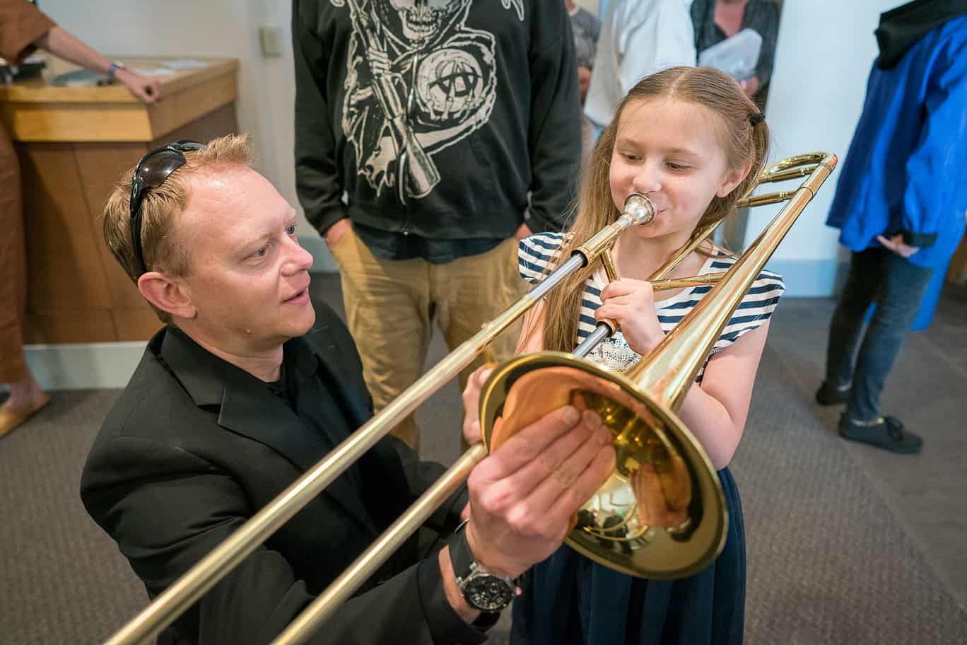 A little girl tries to play a trombone being held by a teacher