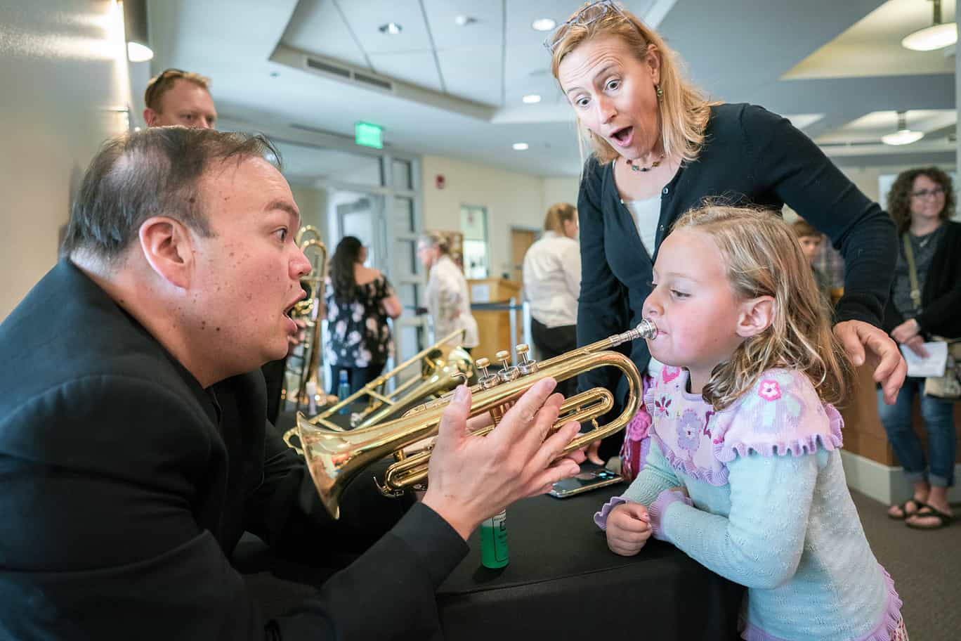 A girl blows into a trumpet held by a teacher, and a woman looks surprised behind her