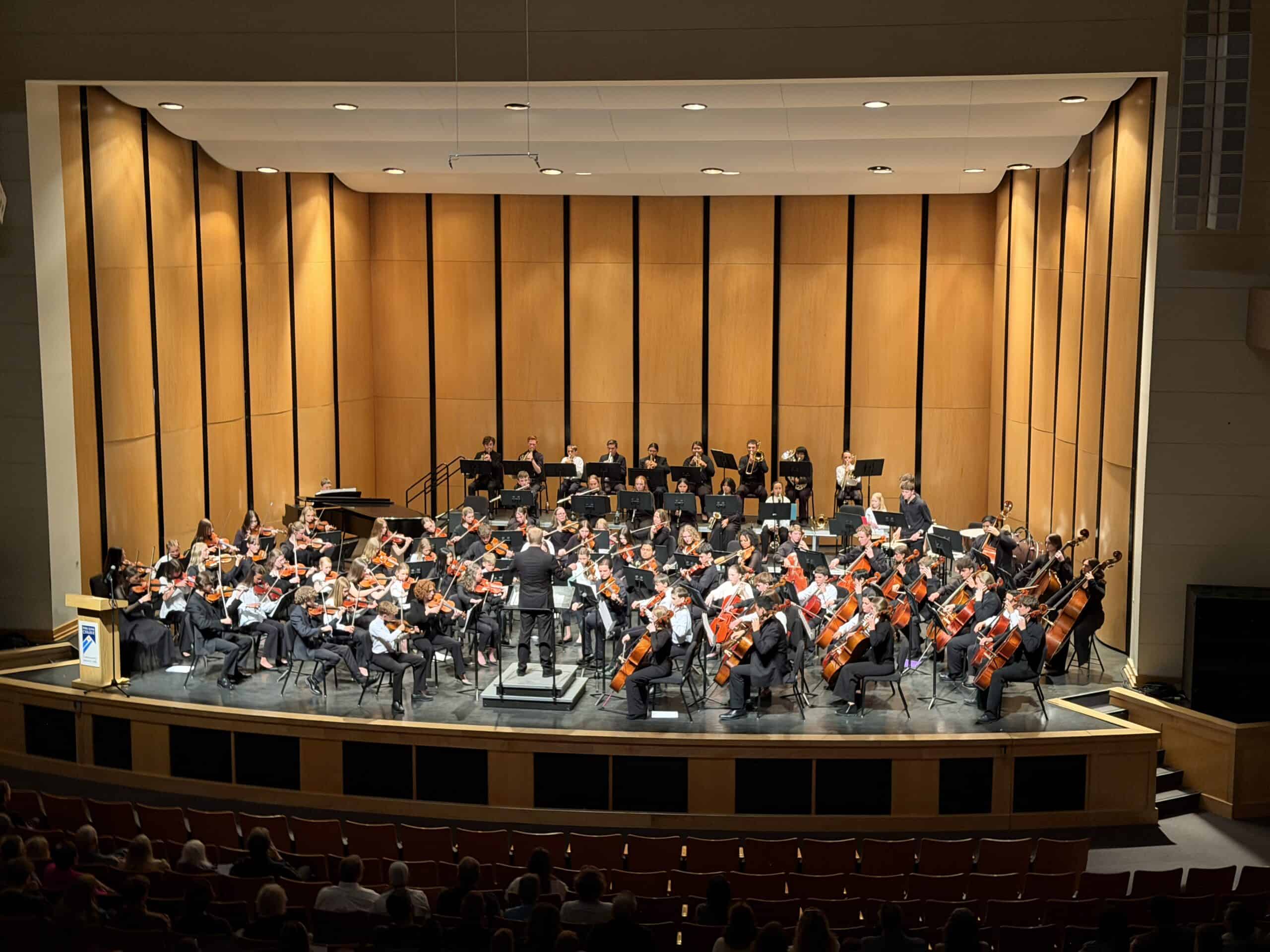 Full orchestra on a wood-paneled concert hall stage, conductor at center, musicians with strings and brass, audience in the foreground.